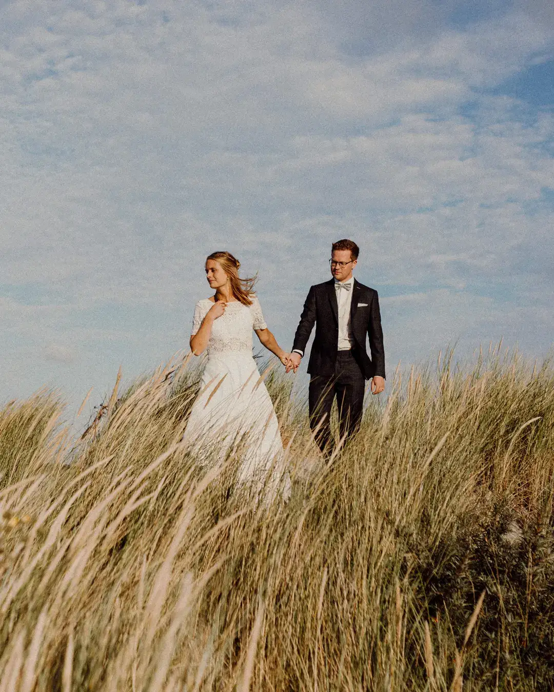 Braut und Bräutigam Hand in Hand in einer Düne auf Ameland mit hohem Gras, mit blauem Himmel im Hintergrund.