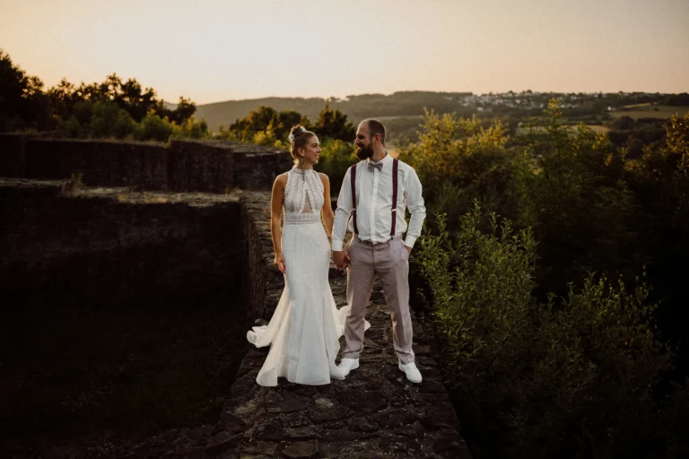 Ein Brautpaar in eleganter Hochzeitskleidung steht auf einer alten Steinmauer von Burg Blankenstein bei Sonnenuntergang, umgeben von grüner Natur. Im Hintergrund sind die Hügel und Landschaft in Hattingen zu sehen.