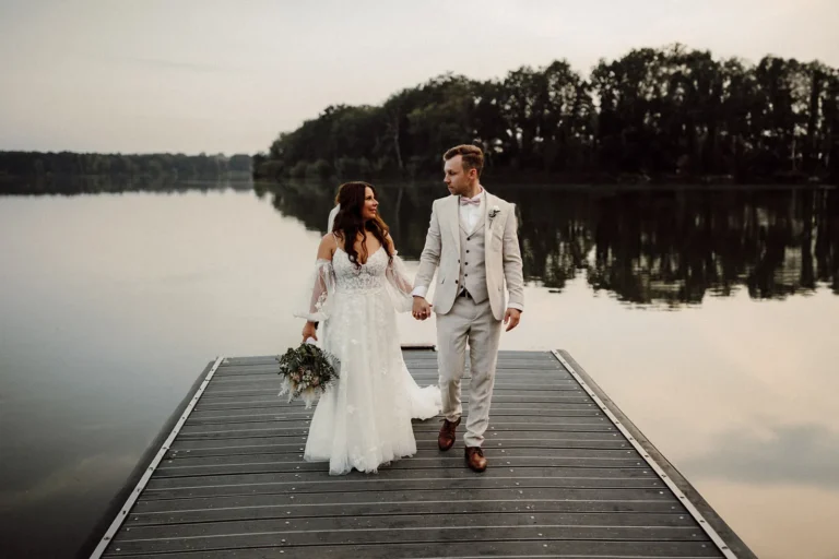 Brautpaar auf einem Steg mit Seeblick in Haltern während ihrer Hochzeit. Die Braut trägt ein elegantes weißes Kleid und der Bräutigam einen stilvollen hellen Anzug. Sie halten sich an den Händen, während der ruhige See und die bewaldete Uferlandschaft den Hintergrund bilden.